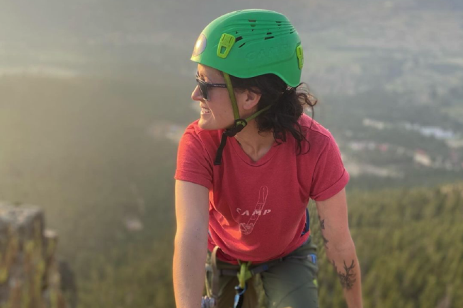 Woman rock climbing in Estes Park with red shirt and green helmet looking off into the distance with valley view behind her